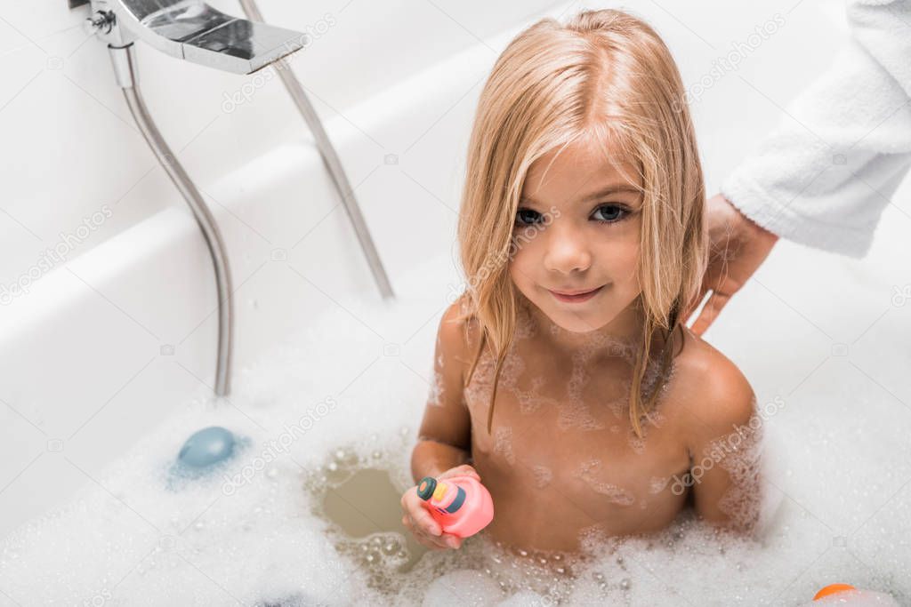 overhead view of cute child taking bath and holding rubber toy near mother 