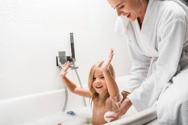 Neşeli anne, elleri başının üstünde banyo köpüğüyle banyo yapan çocuğun yanında. 