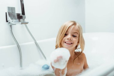 Mutlu çıplak çocuk banyo yaparken banyo köpüğüne bakıyor. 