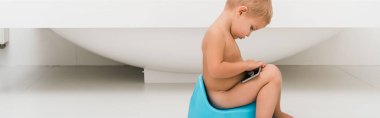 panoramic shot of toddler boy sitting on blue potty and holding smartphone near bathtub  clipart