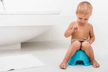 cute toddler boy sitting on blue potty and using smartphone near bathtub  clipart