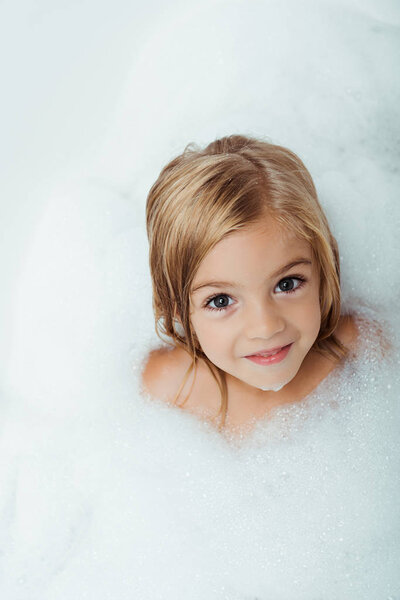 overhead view of cute kid taking bath and looking at camera at home 