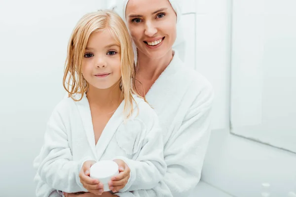 Glückliches Kind Hält Behälter Mit Kosmetikcreme Neben Fröhlicher Mutter Bademantel — Stockfoto