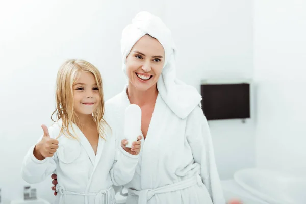 Cheerful Kid Holding White Bottle Showing Thumb Mother Towel Bathrobe — Stock Photo, Image