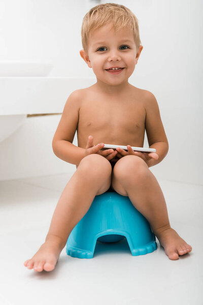 happy toddler boy sitting on blue potty and holding smartphone near bathtub 