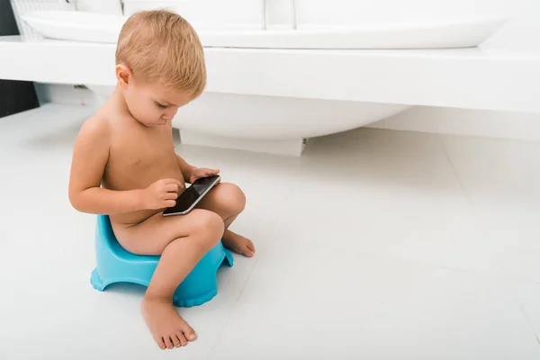 可爱的蹒跚学步的男孩坐在蓝色便盆上 在浴缸附近用智能手机 — 图库照片