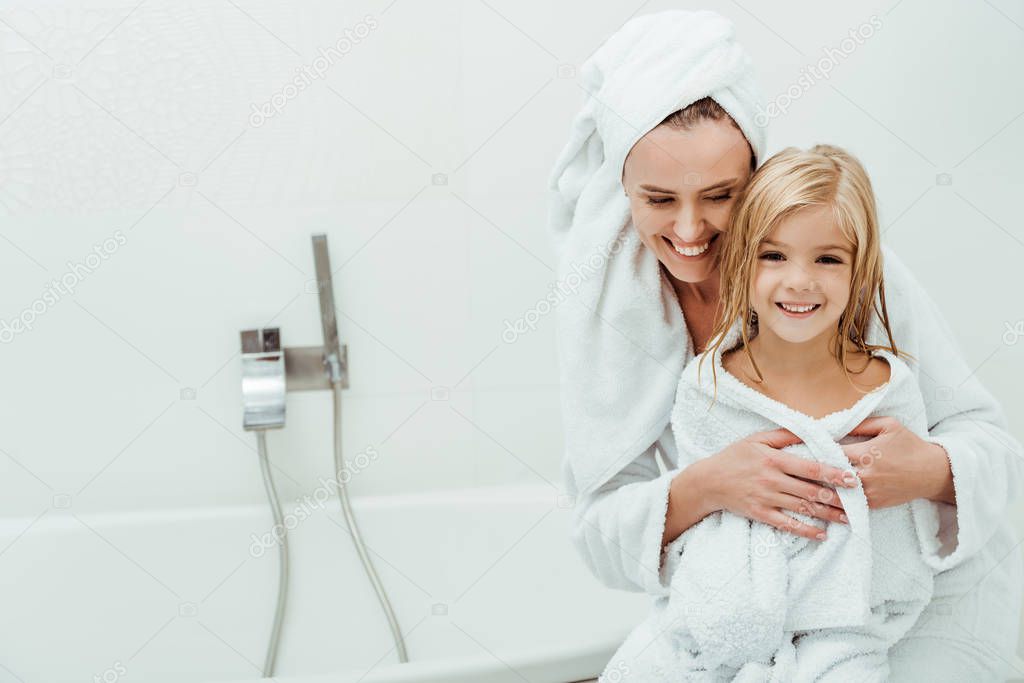 happy mother smiling while hugging cute daughter in bathrobe 