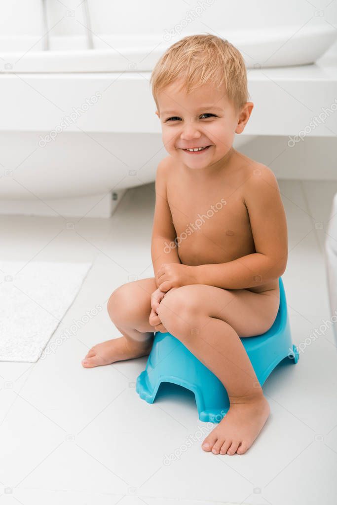 smiling toddler boy sitting on blue potty near bathtub 