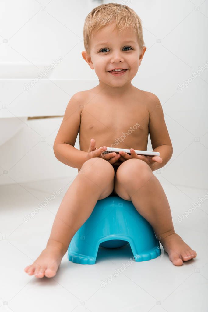 happy toddler boy sitting on blue potty and holding smartphone near bathtub 