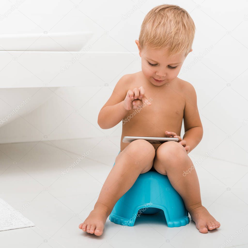 cute toddler boy sitting on potty and using smartphone near bathtub 