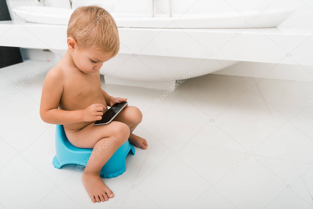 adorable toddler boy sitting on blue potty and using smartphone near bathtub 