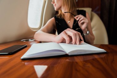 cropped view of businesswoman holding pen near notebook and smartphone with blank screen in private jet  clipart