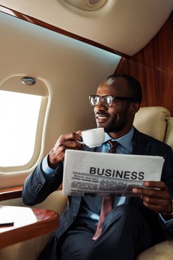 happy african american businessman in glasses holding business newspaper and cup in private plane  clipart