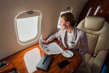 Neşeli iş kadını elinde fincanla akıllı telefonun yanında özel uçakta boş ekranlı bir gazete tutuyor. 