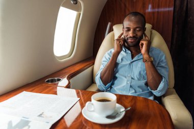 Seçici odak noktası, mutlu Afro-Amerikan bir adamın özel uçakta bardağın yanında içkili müzik dinlemesi. 