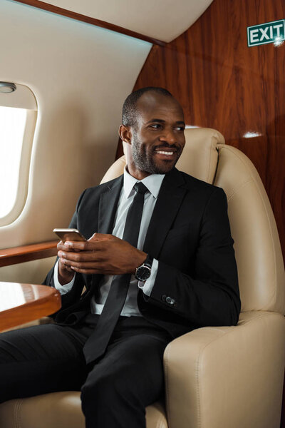 cheerful african american businessman in suit using smartphone in private jet 