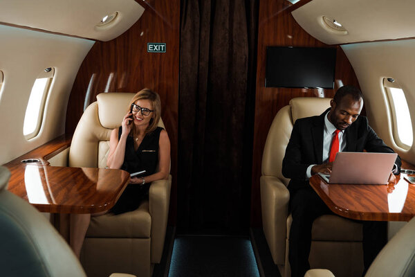 Африканский американский бизнесмен, использующий ноутбук рядом с бизнесвумен разговаривает на смартфоне в частном самолете
 