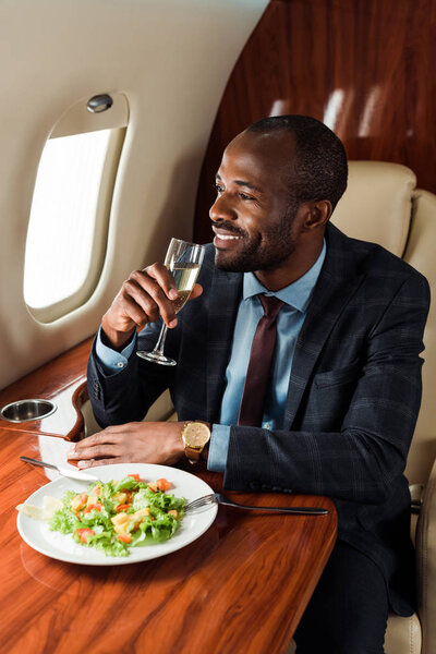 happy african american man with champagne glass near salad in private jet 