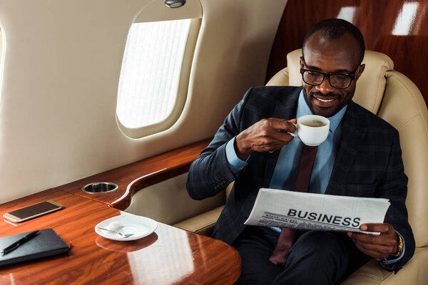 cheerful african american businessman in glasses holding business newspaper and cup in private plane 