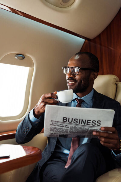 happy african american businessman in glasses holding business newspaper and cup in private plane 