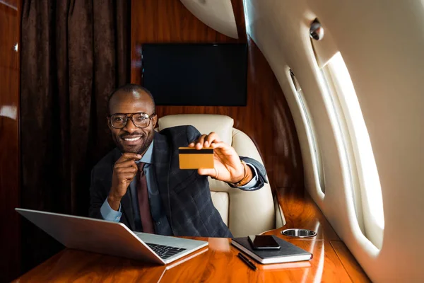 Szczęśliwy Afrykański Biznesmen Trzyma Kartę Kredytową Pobliżu Laptopa Smartfona Samolocie — Zdjęcie stockowe