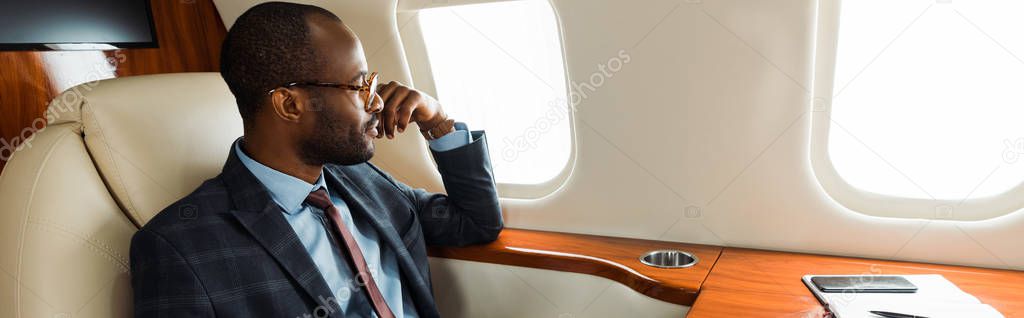 panoramic shot of handsome african american businessman in glasses sitting in private plane 