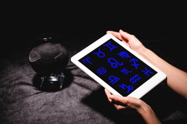 Astrologun elinde dijital tablet, kristal kürenin yanında siyah kadife kumaş üzerinde zodyak tabelaları tutarken çekilmiş görüntüsü.