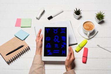 Ofis malzemeleri ve kahve ile birlikte işyerinde elinde zodyak tabelaları olan dijital tablet tutan bir kadın görüntüsü.