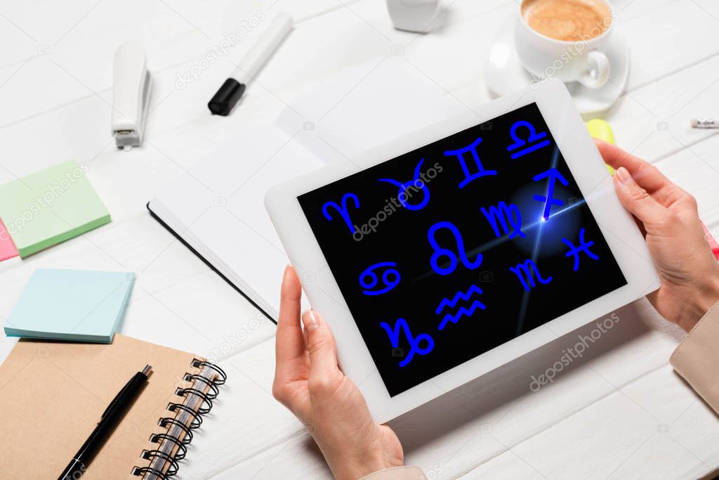 cropped view of woman holding digital tablet with zodiac signs at workplace with office supplies and coffee