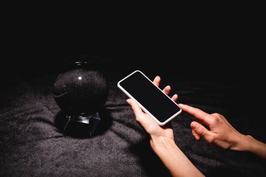 Siyah kadife kumaş üzerinde kristal kürenin yanında akıllı telefon tutan astrolog görüntüsü.