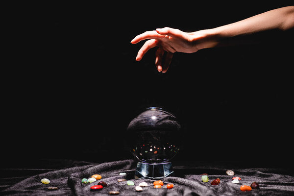 cropped view of fortuneteller hand above crystal ball with fortune telling stones on black velvet cloth isolated on black