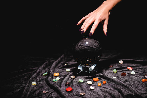 cropped view of fortuneteller hand above crystal ball with fortune telling stones on black velvet cloth isolated on black