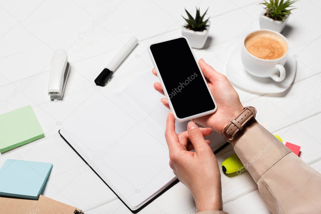 cropped view of woman holding smartphone at workplace with office supplies and coffee