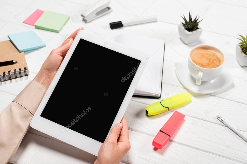 cropped view of woman holding digital tablet at workplace with office supplies and coffee