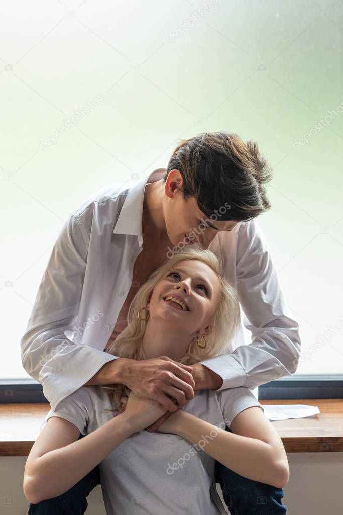 Handsome man hugging smiling girlfriend while sitting on windowsill 
