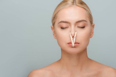 attractive and naked young woman with wooden pin on nose and closed eyes isolated on grey  clipart