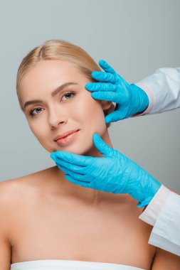 cropped view of beautician in blue latex gloves touching face of young woman isolated on grey  clipart