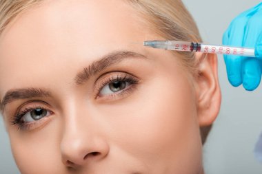 Plastik eldivenli güzellik uzmanının, gri renkli genç bir kadının yanında elinde şırınga tutarken çekilmiş görüntüsü. 