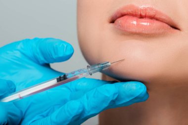 Lastik eldivenli bir güzellik uzmanının elinde gri çeneli bir kızın çenesinin yanında şırınga tutarken görüntüsü. 