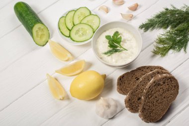 Tasty tzatziki sauce with fresh ingredients and bread on white wooden background clipart