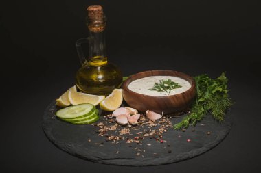 Tzatziki soslu kase, siyah arka planda malzeme, baharat ve zeytinyağı.