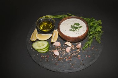 Baharatlı lezzetli tzatziki sosu, zeytinyağı ve siyah arka plandaki koyu tahtadaki malzemeler.