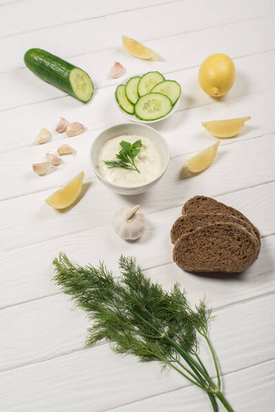 Homemade tzatziki sauce with ingredients, greens and bread on white wooden background