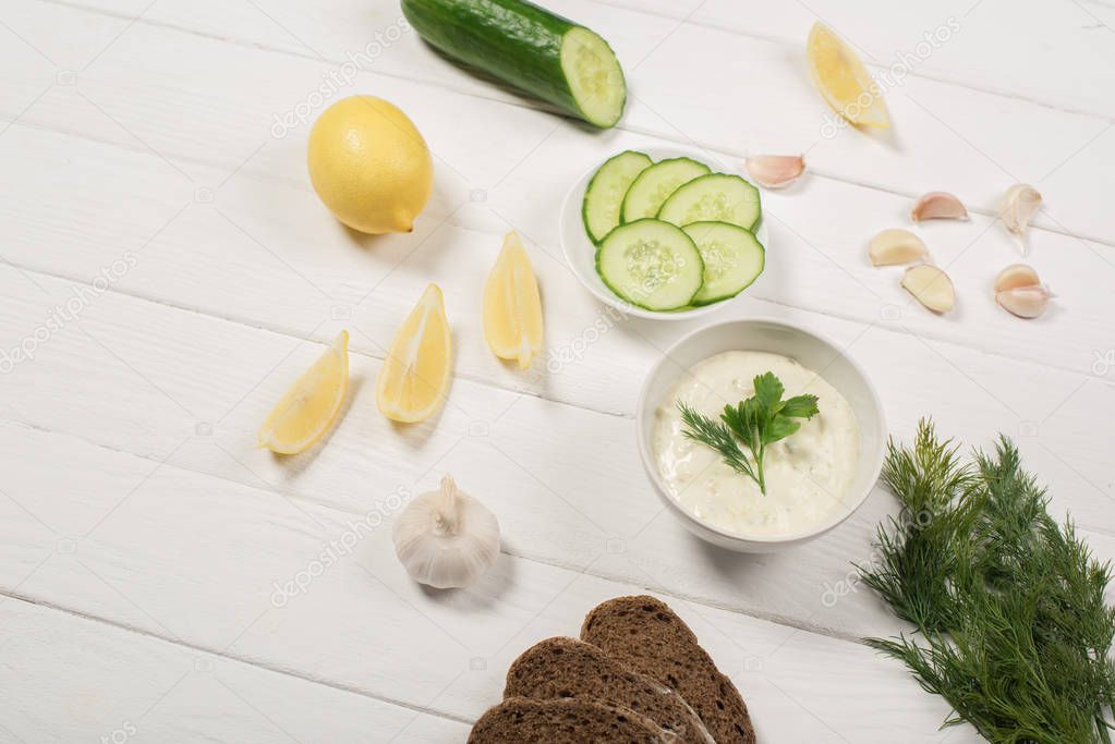 Tzatziki sauce in bowl with fresh ingredients and bread on white wooden background