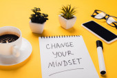 notebook változó gondolkodásmóddal felirat mellett kávéscsésze, cserepes növények, filctoll, okostelefon és szemüveg sárga asztalon