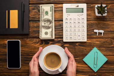 Akıllı telefonun yanında kahve fincanı tutan iş kadını görüntüsü, dolar banknotları, hesap makinesi, kablosuz kulaklıklar ve ahşap masadaki kırtasiye malzemeleri.