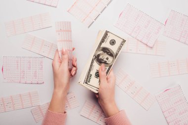 Bir kadının elinde dolar banknotları ve piyango bileti olan beyaz masa üzerinde dağınık piyango kartları olan bir kadın.