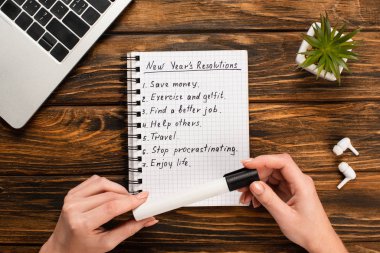 cropped view of businesswoman holding felt-tip pen near notebook with list of new years resolutions near laptop, potted plant and wireless earphones on wooden desk clipart