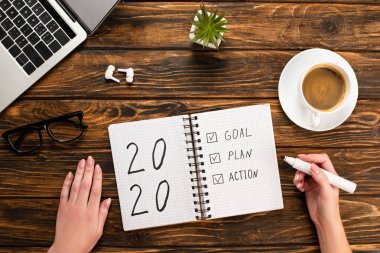 cropped view of businesswoman holding felt-tip pen near notebook with 2020, goal, plan, action lettering near laptop, wireless earphones, coffee cup on wooden desk clipart