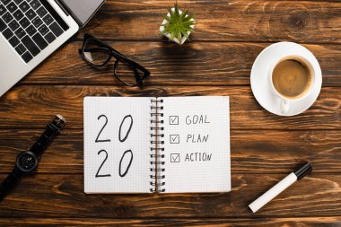 notebook with 2020, goal, plan, action lettering, laptop, coffee cup, felt-tip pen, glasses, wristwatch and plant on wooden desk clipart